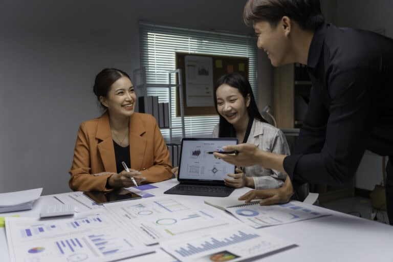 Three young Asian entrepreneurs are smiling while working together, analyzing financial data on a tablet and paperwork during a productive meeting in a modern office